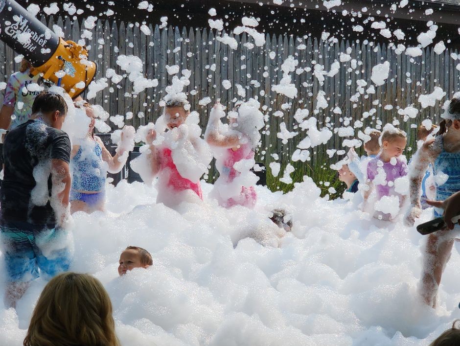 Children playing in foam outdoors with a fence in the background and a foam machine visible on the left.