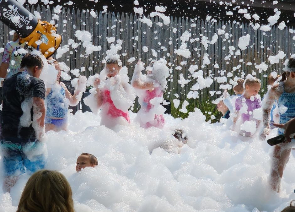 Children playing in foam outdoors with a fence in the background and a foam machine visible on the left.